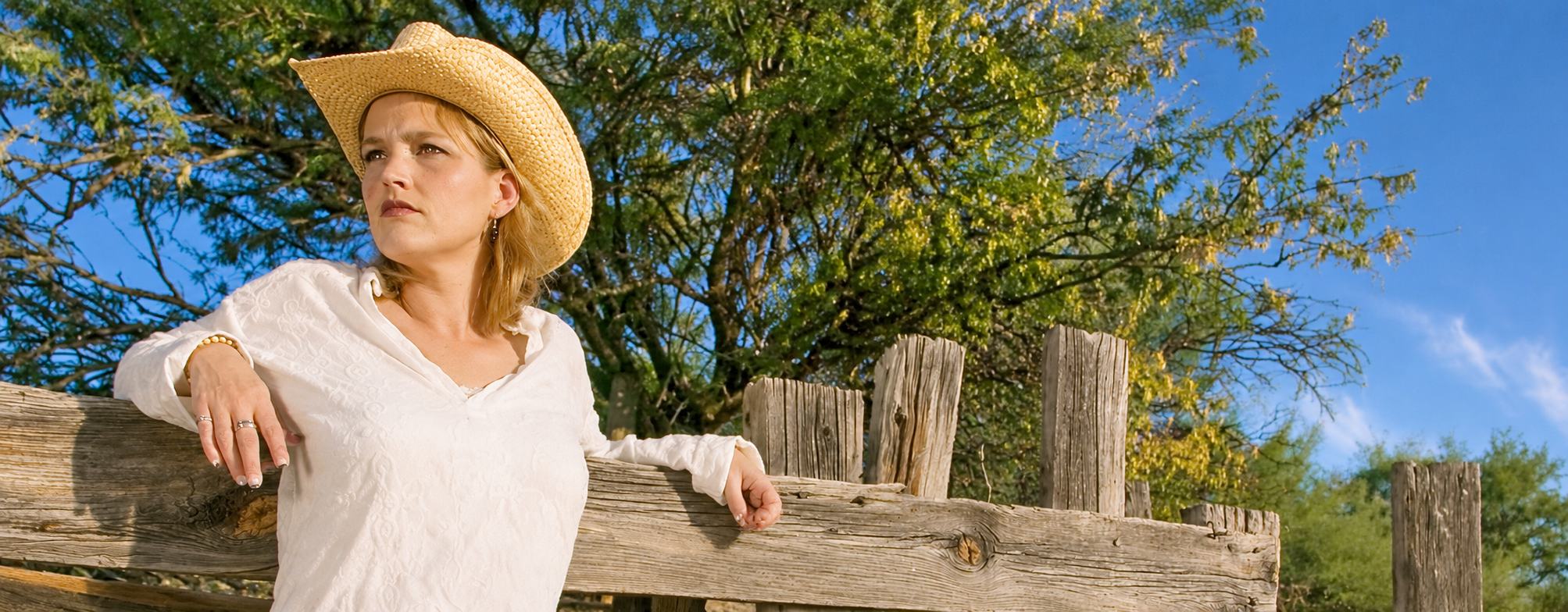 A woman wearing a straw western hat beside a weathered fence on a sunny day